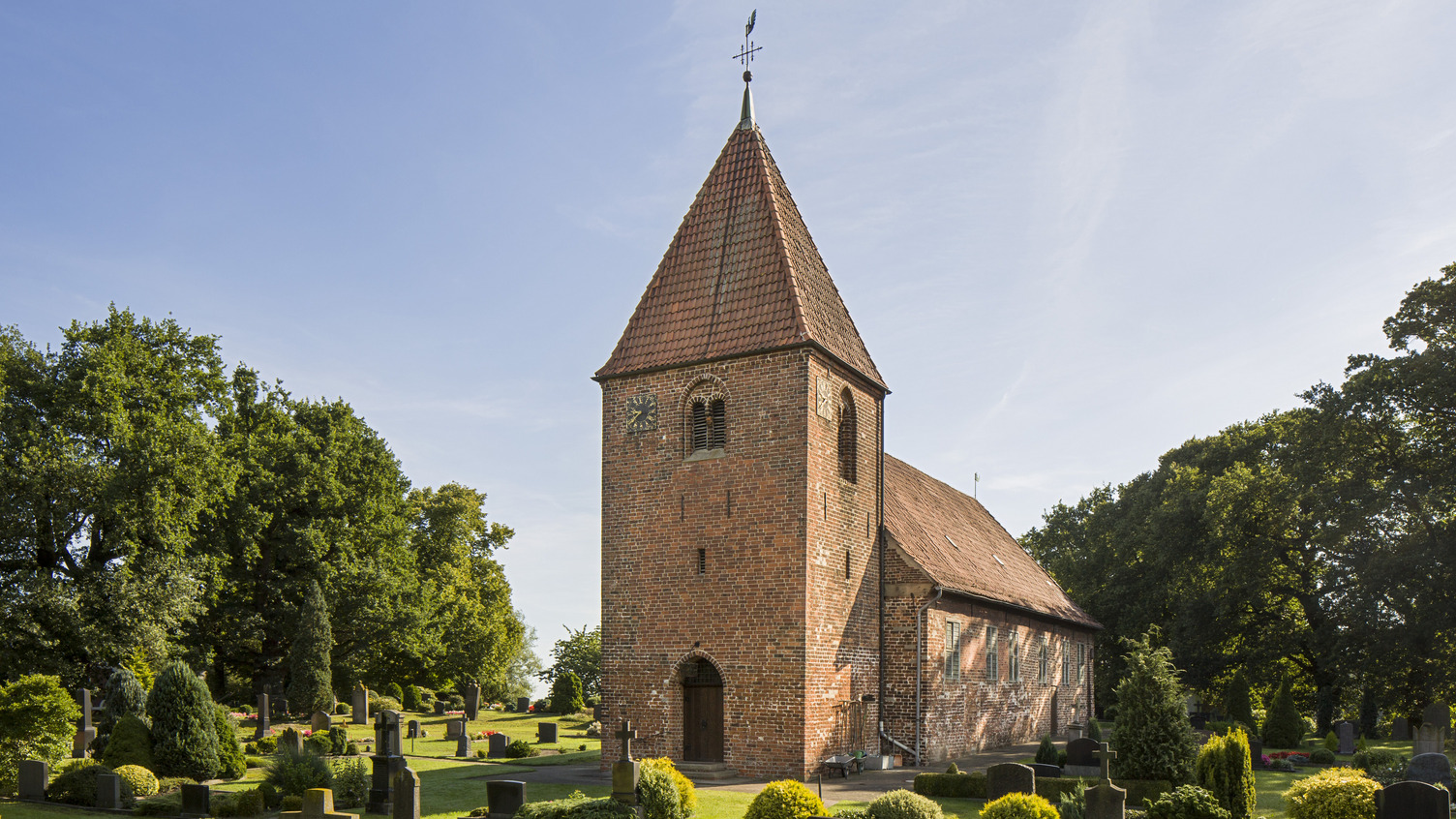 Eine kleine, historische Kirche mit einem hohen, spitzen Dach steht in einem gepflegten Friedhof. Umgeben von grünen Wiesen, Sträuchern und Grabsteinen, strahlt die Szene Ruhe und Würde aus. Der Himmel ist klar und blau, während große Bäume im Hintergrund Schatten spenden.