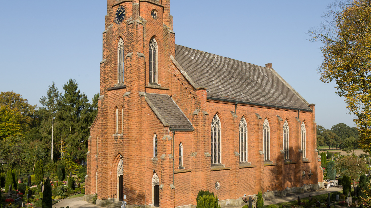 Die Abbildung zeigt eine neugotische Kirche aus roten Ziegelsteinen, umgeben von einem Friedhof. Der hohe Turm mit Uhr und große Fenster prägen die Fassade, während Bäume und Grabstätten im Vordergrund sichtbar sind. Der Himmel ist blau und klar.