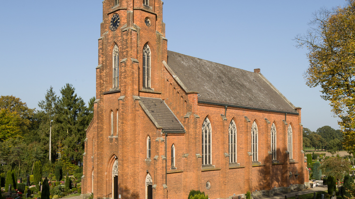 Die Abbildung zeigt eine neugotische Kirche aus roten Ziegelsteinen, umgeben von einem Friedhof. Der hohe Turm mit Uhr und große Fenster prägen die Fassade, während Bäume und Grabstätten im Vordergrund sichtbar sind. Der Himmel ist blau und klar.