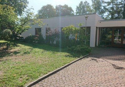 Ein modernes, einstöckiges Gebäude mit großen Fenstern und einer begrünten Umgebung. Der Pflasterweg führt zur Eingangstür, umgeben von Bäumen und Sträuchern. 