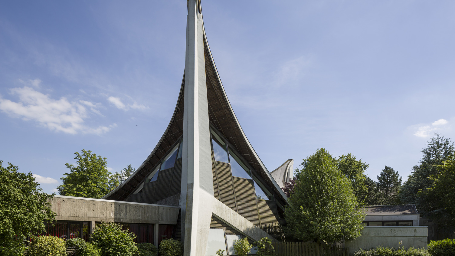 Das Bild zeigt ein modernes Gebäude mit einer charakteristischen geschwungenen Dachkonstruktion und einer hohen, schrägen Wand. Umgeben von Bäumen und Sträuchern, steht das Gebäude in einer grünen Umgebung unter einem klaren blauen Himmel.