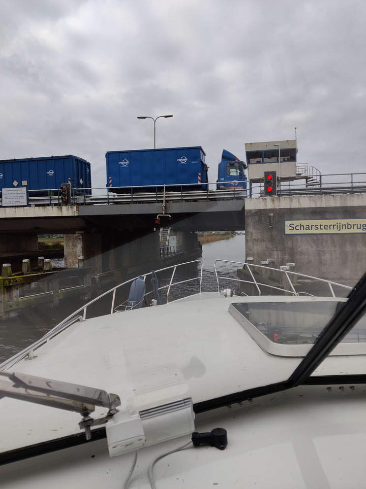 Scharsterrijnbrug Eine Sicht auf den Bug eines Bootes und auf eine Brücke vor dem Boot, unter der sich das Wasser befindet. Ein Lkw mit blauen Containern fährt über die Brücke. Zwei rote Lichter signalisieren, dass die Brücke nicht passiert werden kann.