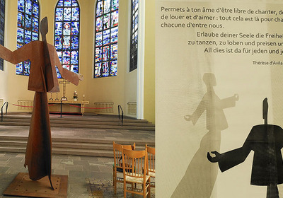Eine große, abstrakte Skulptur eines aufrechten Menschen mit erhobenen Armen steht in einem Kirchenraum. Im Hintergrund befinden sich bunte Fenster. Neben der Skulptur ist ein Zitat zu sehen, das zur Freiheit des Singens, Tanzens und Liebens einlädt.