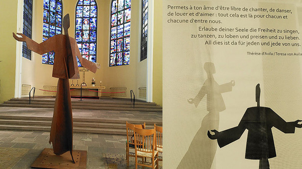 Eine große, abstrakte Skulptur eines aufrechten Menschen mit erhobenen Armen steht in einem Kirchenraum. Im Hintergrund befinden sich bunte Fenster. Neben der Skulptur ist ein Zitat zu sehen, das zur Freiheit des Singens, Tanzens und Liebens einlädt.