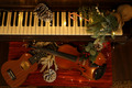 Ein Klavier mit Tasten ist im Hintergrund sichtbar, während im Vordergrund eine Violine und eine Ukulele auf einem roten Untergrund liegen. Um die Instrumente sind weihnachtliche Dekorationen wie Tannenzweige, Zapfen und Lichterketten arrangiert, die eine festliche Atmosphäre schaffen.