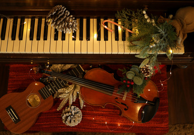 Ein Klavier mit Tasten ist im Hintergrund sichtbar, während im Vordergrund eine Violine und eine Ukulele auf einem roten Untergrund liegen. Um die Instrumente sind weihnachtliche Dekorationen wie Tannenzweige, Zapfen und Lichterketten arrangiert, die eine festliche Atmosphäre schaffen.