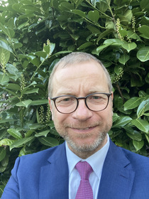 Ein lächelnder Mann mit Brille trägt einen blauen Anzug und eine pinke Krawatte. Er steht vor einem dichten, grünen Laub.
