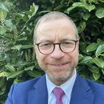 Ein lächelnder Mann mit Brille trägt einen blauen Anzug und eine pinke Krawatte. Er steht vor einem dichten, grünen Laub.