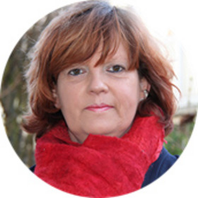 Porträt einer Frau mit schulterlangen, rötlichen Haaren, die einen roten Schal trägt. Sie steht vor einem unscharfen Hintergrund. Ihr Gesichtsausdruck ist neutral und freundlich.