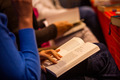 Eine Person hält ein aufgeschlagenes Buch in den Händen. Die Seiten sind beschriftet und die Person scheint zu lesen. Im Hintergrund sind unscharfe Silhouetten weiterer Personen, die ebenfalls Bücher halten.