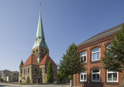 Die Ansicht zeigt eine steinerne Kirche mit spitzem, kupferfarbenem Dach und einem schönen, detaillierten Kirchturm. Neben der Kirche steht ein rotes Backsteingebäude. Der Himmel ist klar und blau.