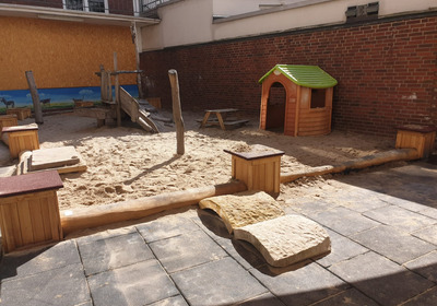 Das Bild zeigt einen Spielplatz mit einem Sandkasten in der Mitte, einer Rutsche  und einer kleinen Holzhütte mit grünem Dach. Um den Sandkasten stehen Holzbänke, und der Boden ist mit weichen Platten ausgelegt. 