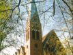 Eine Kirche mit einem hohen, spitzen Turm und einer markanten Fassade aus roten Ziegeln, umgeben von frischem Laub. Der Himmel ist klar, und die Sonnenstrahlen beleuchten die Bäume, die die Kirche teilweise umrahmen.