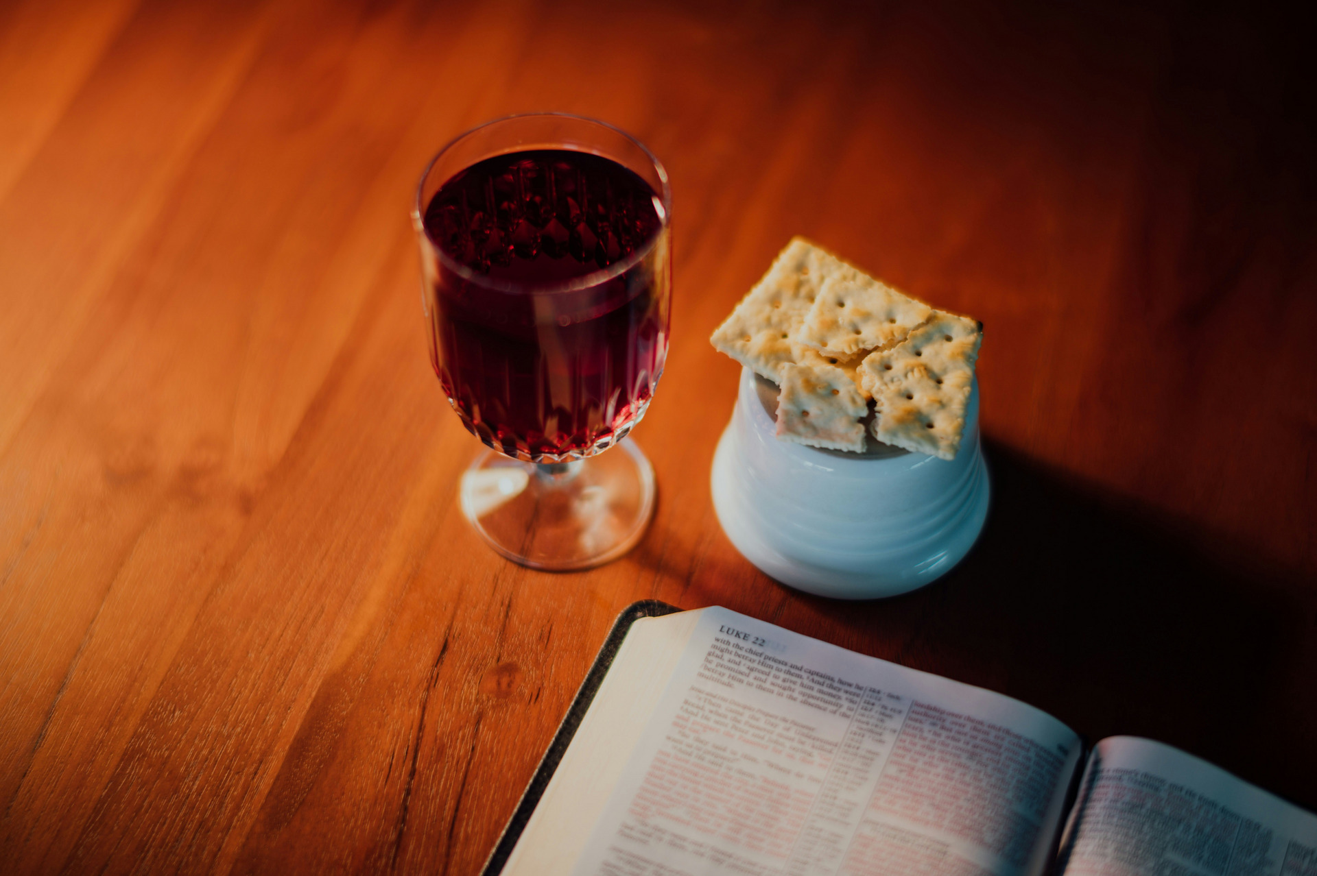 Auf einem Holztisch liegen ein geöffnetes Buch, ein Glas mit rotem Getränk und eine kleine Schüssel mit Crackern. Das Bild vermittelt eine Atmosphäre der Besinnung oder des gemeinsamen Essens.