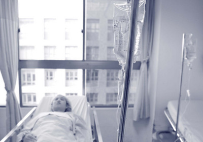Ein Person liegt starr in einem Krankhausbett, im Vordergrund steht ein Infusionsständer.