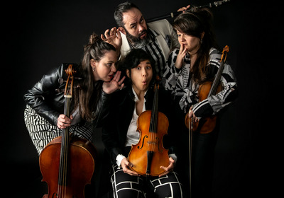 Eine Gruppe von vier Musikern posiert kreativ vor einem schwarzen Hintergrund. Eine Person hält eine Violine, während die anderen schüchterne Gesten machen und leise miteinander kommunizieren. Die Musiker tragen unterschiedliche, auffällige Kleidung.