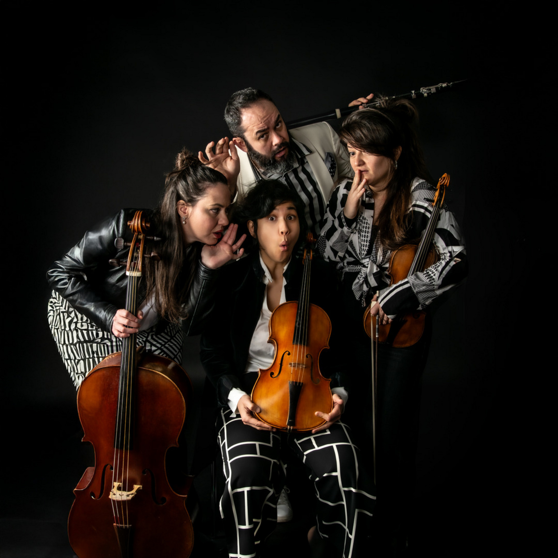 Eine Gruppe von vier Musikern posiert kreativ vor einem schwarzen Hintergrund. Eine Person hält eine Violine, während die anderen schüchterne Gesten machen und leise miteinander kommunizieren. Die Musiker tragen unterschiedliche, auffällige Kleidung.