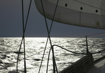 Ein Segelboot fährt über glitzerndes Wasser, das das Licht reflektiert. Im Hintergrund ist der Himmel bewölkt. Die Segel sind aufgebläht und die Takelage ist sichtbar, während das Boot sanft durch die Wellen gleitet.
