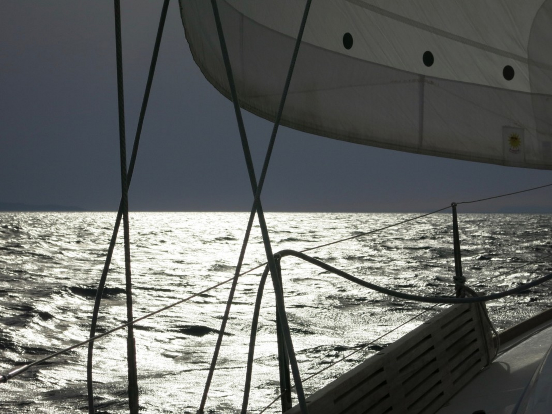 Ein Segelboot fährt über glitzerndes Wasser, das das Licht reflektiert. Im Hintergrund ist der Himmel bewölkt. Die Segel sind aufgebläht und die Takelage ist sichtbar, während das Boot sanft durch die Wellen gleitet.