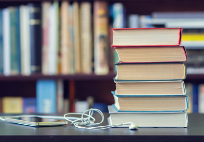 Das Foto zeigt einen Tisch mit einem Bücherstapel und einem Smartphone mit Kopfhörern. Im Hintergrund eine Bücherregal.