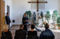 Ein Trauergottesdienst in einer Kirche. Gäste Altar mit Kreuz und eine Pfarrerin sind zu sehen.