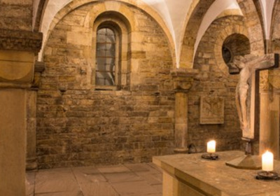 Ein historischer Raum mit steinernen Wänden und gewölbter Decke. In der Mitte steht ein Altar mit einer Christusfigur, die am Kreuz hängt. Auf dem Altar brennen zwei Kerzen, die eine feierliche Stimmung erzeugen.