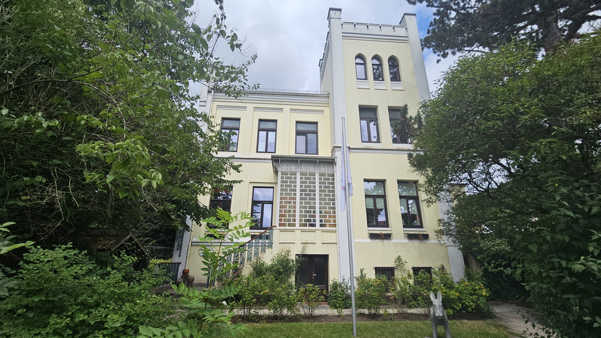 Ein mehrstöckiges gelbes Gebäude mit großen Fenstern und einem dekorativen, grafischen Element an der Fassade. Umgeben von üppigem Grün und Bäumen, bietet die Umgebung einen ruhigen und naturverbundenen Eindruck. Ein geschmackvoller Garten vervollständigt das Bild.