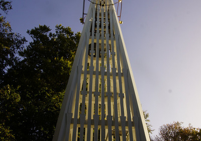 Ein hoher, schmaler Turm aus weißem Holz, der in die Höhe ragt. Der Turm hat vertikale Streben und eine geometrische Spitze. Der Himmel ist blau, und Bäume umgeben die Basis des Turms, was eine ruhige, naturnahe Atmosphäre schafft.