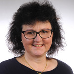 Porträt einer Frau mit kurzen, dunklen Haaren und Brille. Sie trägt ein schwarzes Oberteil und eine goldene Halskette mit einem herzförmigen Anhänger. Die Frau lächelt freundlich in die Kamera. Der Hintergrund ist hell und neutral.