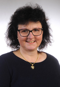 Porträt einer Frau mit kurzen, dunklen Haaren und Brille. Sie trägt ein schwarzes Oberteil und eine goldene Halskette mit einem herzförmigen Anhänger. Die Frau lächelt freundlich in die Kamera. Der Hintergrund ist hell und neutral.