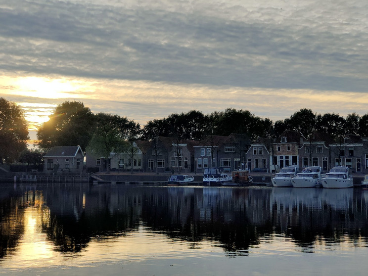 Blockzijl Eine ruhige Wasseroberfläche spiegelt die Farben des Sonnenuntergangs wider. Beidseitig des Wassers sind reizvolle Häuser und Boote zu sehen, umgeben von Bäumen. Der Himmel ist mit sanften Wolken bedeckt, die das Licht der untergehenden Sonne reflektieren.
Rechts im Bild liegen unsere drei Boote nebeneinander vertäut.