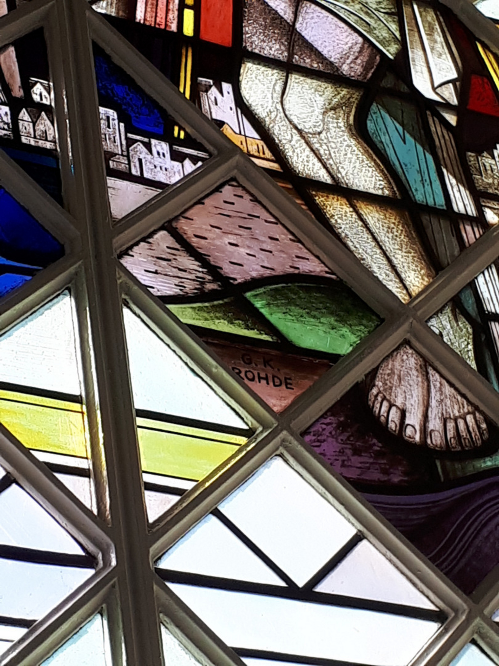 Ein farbenfrohes Kirchenfenster zeigt die Füße einer Figur, umgeben von geometrischen Mustern und Landschaftselementen. Die Szene vermittelt einen spirituellen Eindruck und ist reich an Details, die das Licht kreativ durch die bunten Gläser filtern.