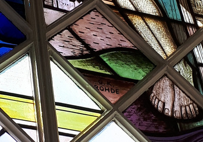 Ein farbenfrohes Kirchenfenster zeigt die Füße einer Figur, umgeben von geometrischen Mustern und Landschaftselementen. Die Szene vermittelt einen spirituellen Eindruck und ist reich an Details, die das Licht kreativ durch die bunten Gläser filtern.