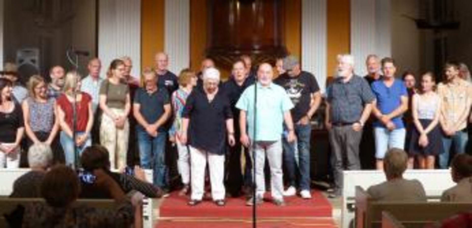 Eine Gruppe von Personen steht auf einer Bühne in einer Kirche. Im Vordergrund stehen zwei Personen, die im Mittelpunkt stehen, während die anderen Mitglieder der Gruppe hinter ihnen stehen und interessiert zuschauen. Die Atmosphäre ist freundlich und musikalisch, möglicherweise bei einer Aufführung oder Veranstaltung.