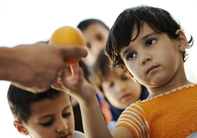 Ein Kind nimmt ein Stück Obst von einem Erwachsenem entgegen. Im Hintergrund sehen weitere Kinder zu.