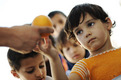 Ein Kind nimmt ein Stück Obst von einem Erwachsenem entgegen. Im Hintergrund sehen weitere Kinder zu.