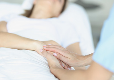 Das Foto zeigt einen nicht erkennbaren Menschen in einem Krankenbett. Jemand legt die Hand auf die Hände der Patientin