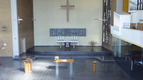 Ein helles Kircheninnere mit einem Altar, auf dem vier Kerzen stehen. Über dem Altar hängt ein großes Wandbild. Ein Kreuz schwebt an der Wand, und im Hintergrund sind Buntglasfenster zu sehen. Es gibt eine Tischdekoration mit Blumen.