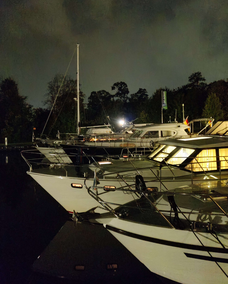 Sneek bei Nacht Mehrere Yachten sind im Hafen bei Nacht zu sehen. Die Boote haben helle Lichter an Deck, die sich im Wasser spiegeln. Der Himmel ist bewölkt, und die Umgebung wirkt ruhig. Der Fokus liegt auf den bereitliegenden Yachten.