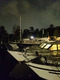Mehrere Yachten sind im Hafen bei Nacht zu sehen. Die Boote haben helle Lichter an Deck, die sich im Wasser spiegeln. Der Himmel ist bewölkt, und die Umgebung wirkt ruhig. Der Fokus liegt auf den bereitliegenden Yachten.