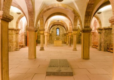 Eine große, historische Krypta mit hohen romanischen Bögen und Säulen. In der Mitte steht ein schlichtes, erhöhtes Altarkreuz, während der Boden mit großen Steinplatten bedeckt ist. Sanftes Licht fällt durch ein Fenster, das die Atmosphäre beruhigend macht.