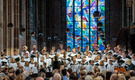 Ein Kinder- und Jugendchor singt in einer Kirche. Die Mitglieder tragen weiße Gewänder mit schwarzen Details und stehen vor einem bunten Glasfenster. Die Zuschauer sind im Hintergrund zu sehen und hören gebannt zu.