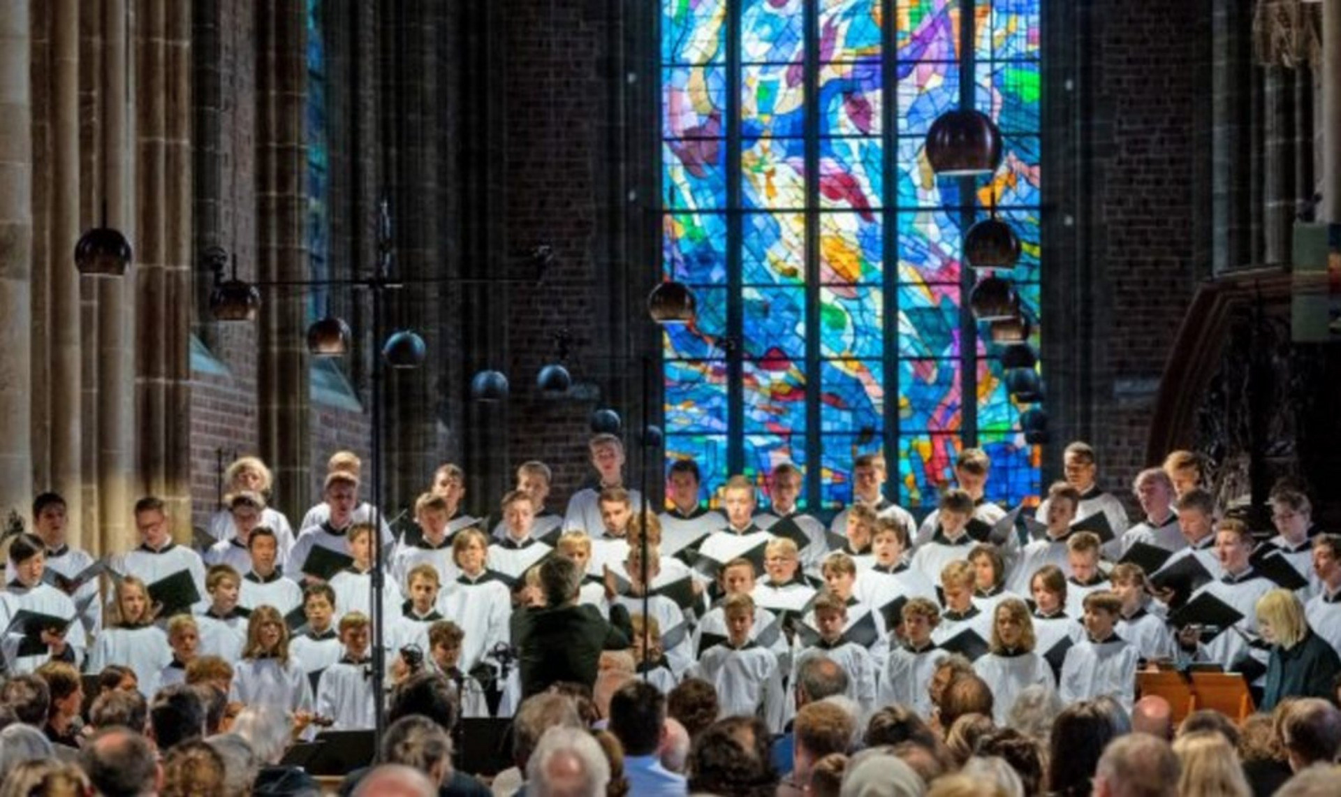 Ein Kinder- und Jugendchor singt in einer Kirche. Die Mitglieder tragen weiße Gewänder mit schwarzen Details und stehen vor einem bunten Glasfenster. Die Zuschauer sind im Hintergrund zu sehen und hören gebannt zu.