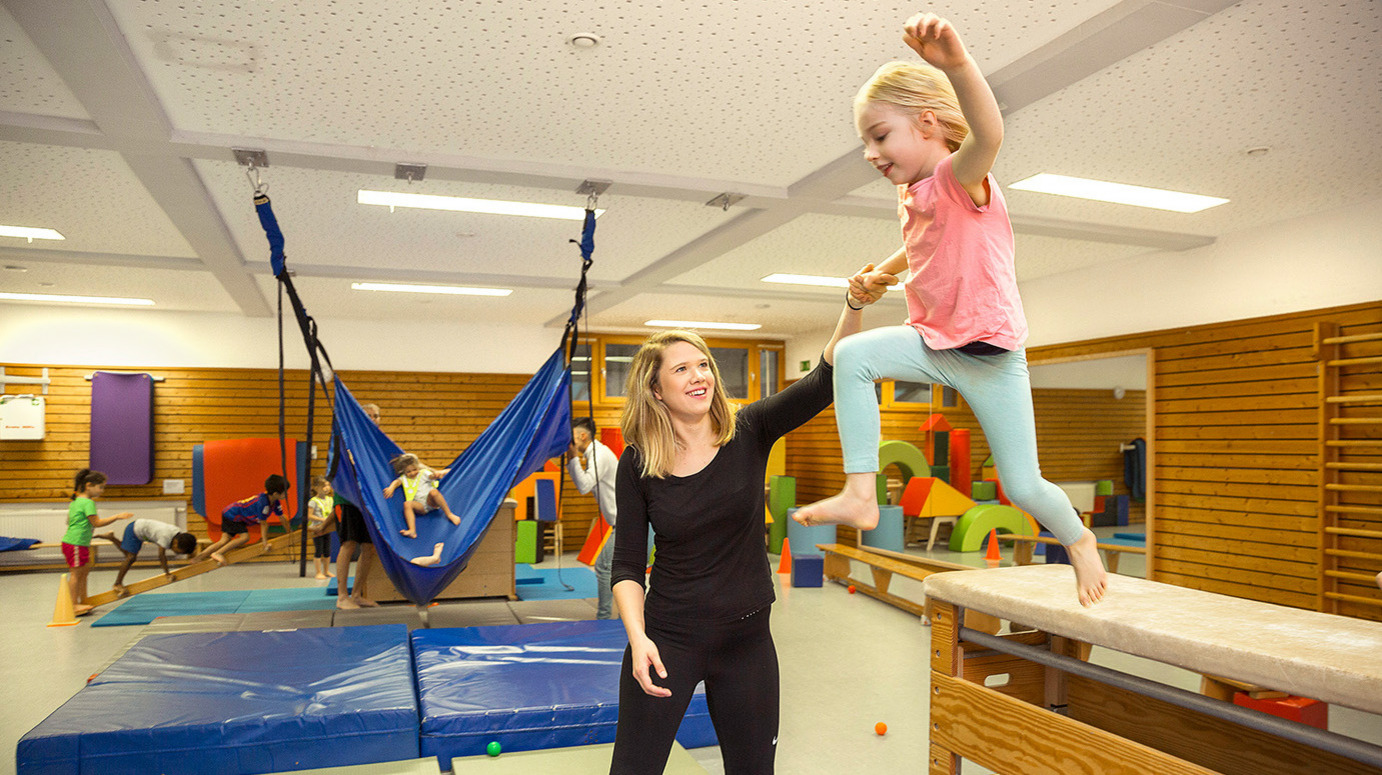 Während einer Sportstunde unterstützt eine Erzieherin ein Kind beim Sprung von einem Turngerät.