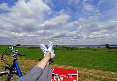 Eine Person sitzt gemütlich und hat ihre Beine auf einem Fahhradsattel ausgestreckt. Sie überblickt eine weite grüne Landschaft mit blauem Himmel und weißen Wolken.
