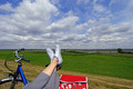 Eine Person sitzt gemütlich und hat ihre Beine auf einem Fahhradsattel ausgestreckt. Sie überblickt eine weite grüne Landschaft mit blauem Himmel und weißen Wolken.
