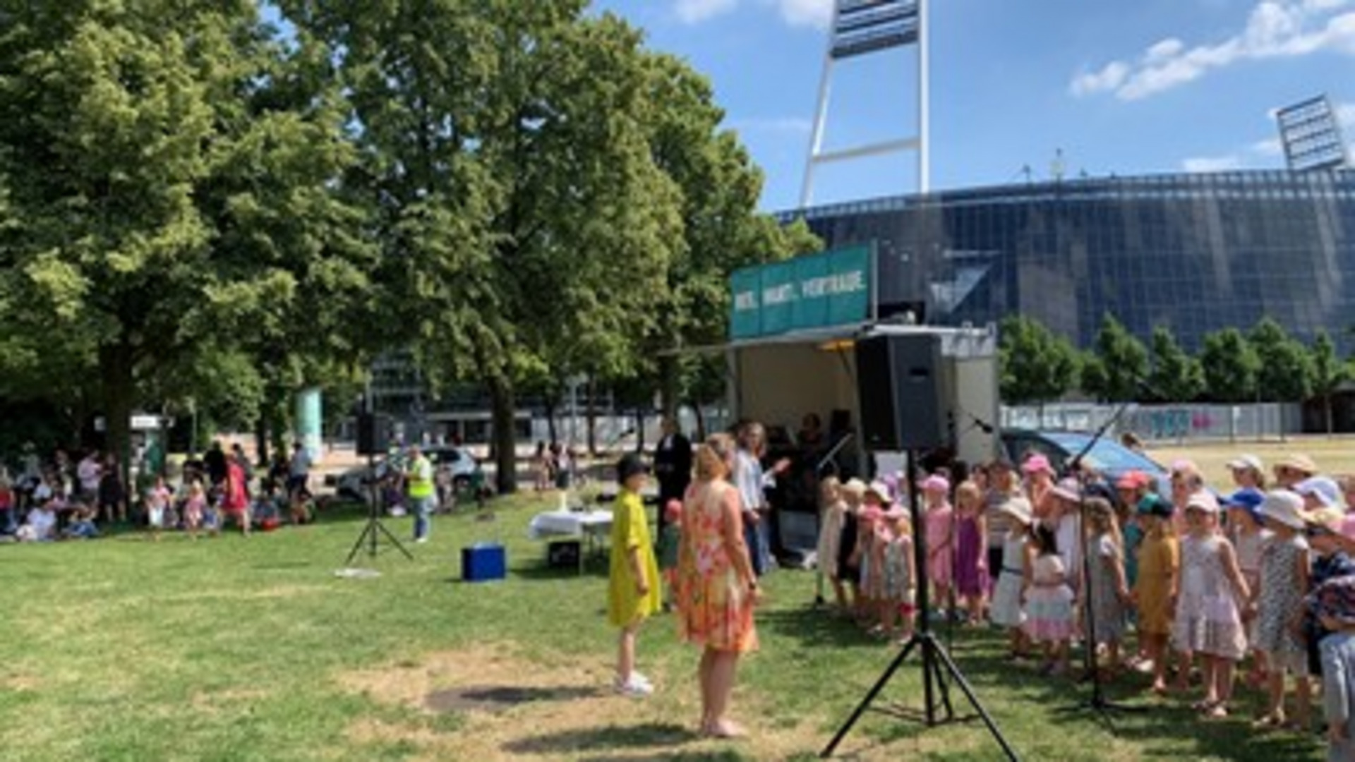 Eine Gruppe von Kindern in bunten Kleidern steht auf einer Wiese und singt. Im Hintergrund ist eine Bühne mit einem Banner zu sehen. Einige Erwachsene stehen davor und beobachten die Aufführung. Bäume umgeben den Bereich, und die Sonne scheint.