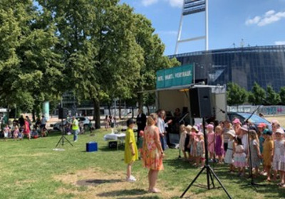 Eine Gruppe von Kindern in bunten Kleidern steht auf einer Wiese und singt. Im Hintergrund ist eine Bühne mit einem Banner zu sehen. Einige Erwachsene stehen davor und beobachten die Aufführung. Bäume umgeben den Bereich, und die Sonne scheint.