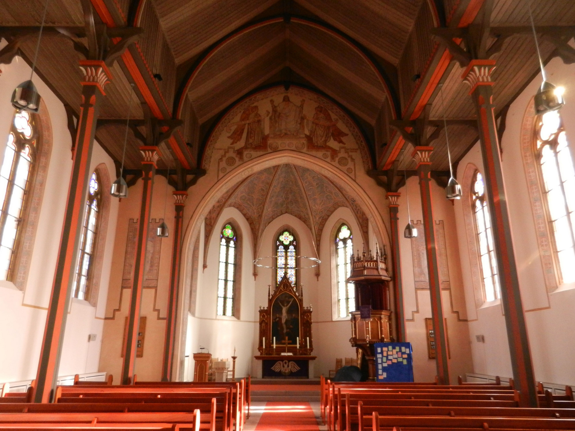 Die Aufnahme zeigt das Innere einer Kirche mit hohen, dekorierten Wänden und einer hohen Decke. In der Mitte befindet sich ein Altar, flankiert von Bänken. Große, bunte Fenster lassen Licht herein, während die gesamte Atmosphäre ruhig und andächtig wirkt.