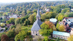 Luftaufnahme einer Kirche mit spitzem Turm, umgeben von bunter Herbstvegetation. Im Vordergrund sind Gräber und eine Wiese sichtbar. Im Hintergrund sind Wohngebäude und Bäume, die die ländliche Umgebung in lebhaften Farben präsentieren.